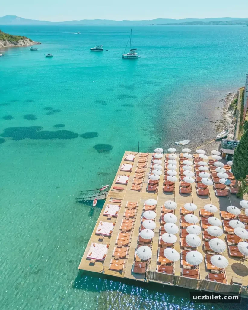 Berrak turkuaz renkteki denize uzanan ahşap iskelede, sıra sıra dizilmiş çok sayıda şezlong ve beyaz şemsiyelerle dolu, uzaklarda yelkenlilerin ve teknelerin yüzdüğü, güneşli bir günde çekilmiş bu görüntü, sakin bir deniz kenarı tatilini çağrıştırıyor.