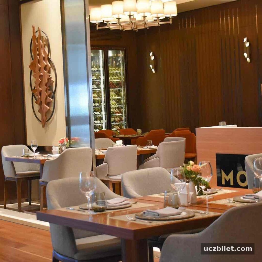 DoubleTree by Hilton Skopje'nin şık Monte Restoranı'nda, özenle düzenlenmiş ahşap masalar, krem renkli sandalyeler, modern avize, duvarda soyut ahşap sanat eseri ve arka planda bir şarap dolabıyla Üsküp seyahatinde konforlu bir gastronomi deneyimi sunan çağdaş ve lüks bir yemek alanı.