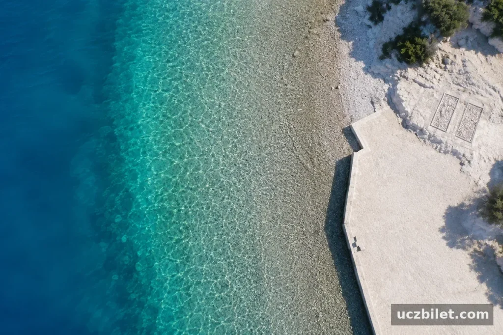 İzmir'de denize girilebilecek en güzel plajlardan biri olan Foça'daki Sazlıca Koyu'nun havadan çekilmiş bu görüntüsünde, berrak turkuaz sularında taşlı deniz tabanı net bir şekilde görülürken, kıyı şeridinde uzanan çakıllı plaj ve yanındaki beton platformla huzurlu bir manzara sunuluyor.