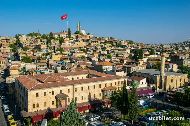 Ön planda kiremit çatılı, taş bir yapı ve hemen yanında minareli bir camiyle, Gaziantep'in yamaçlara yayılan geleneksel evleri ve tepede gururla dalgalanan Türk bayrağı şehrin köklü tarihini ve dokusunu gözler önüne seriyor.