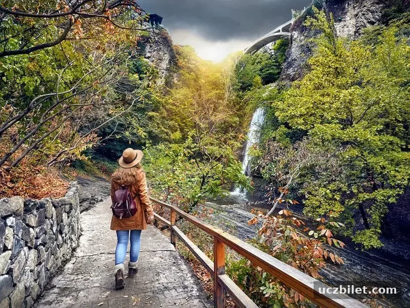 Şapkalı bir turist kadın, Gürcistan'ın Tiflis şehrinde, bulutlu bir havada, sonbahar ağaçlarının bulunduğu botanik bahçesindeki şelaleye doğru gidiyor.