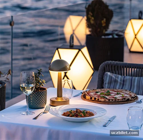 Su kenarında şık bir akşam yemeği masası; üzerinde makarna, pizza, beyaz şarap kadehi ve loş ışıklı bir masa lambası bulunurken, arka planda aydınlatılmış geometrik fenerler ve karanlık deniz romantik bir atmosfer yaratıyor.
