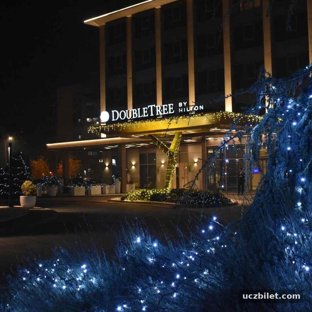 Üsküp seyahat rehberi kapsamında konaklama seçeneklerinden biri olan DoubleTree by Hilton Skopje otelinin gece çekilmiş, girişindeki parlak sarı ve beyaz ışıklarla aydınlanmış tabelası ve süslü çalılarla davetkar görünen ön cephesi.