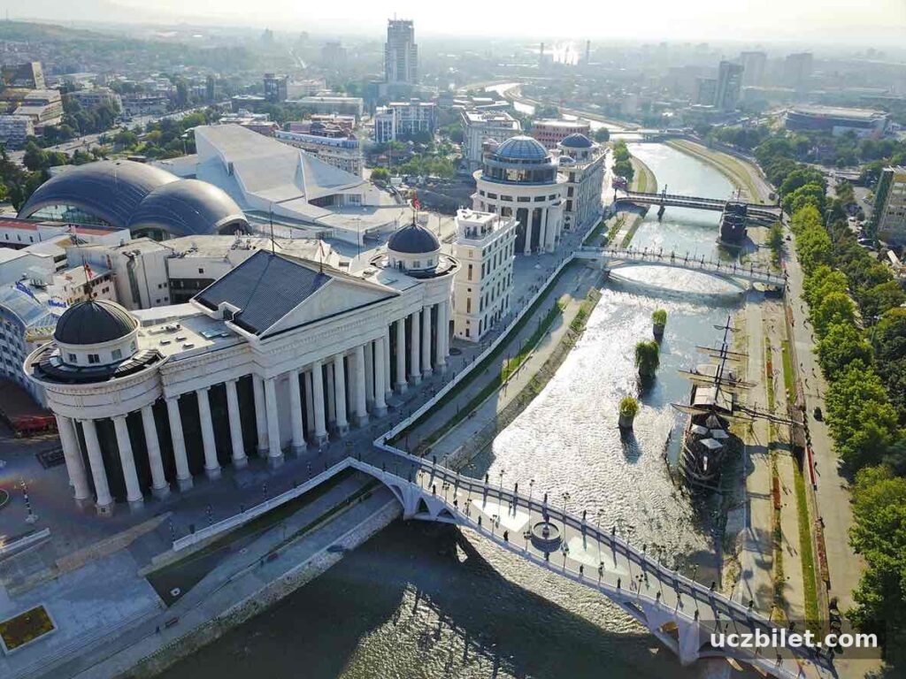 Üsküp'ün merkezinden geçen Vardar Nehri'nin kuşbakışı görünümünde, modernizasyon projeleriyle inşa edilmiş, sütunlu ve kubbeli gösterişli yapılar, heykellerle bezeli köprüler ve nehirdeki gemi replikaları, şehrin zengin ve katmanlı tarihinden kalan izleriyle bugünkü karmaşık yapısını gözler önüne seriyor.
